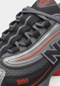 Baskets New Balance 1000 gris et noires avec des accents orange et des bandes réfléchissantes sur la languette et le panneau latéral.
