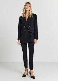 Blazer nero doppiopetto con ampi rever, texture del tessuto e tasche frontali, abbinato a pantaloni neri slim-fit e tacchi a punta neri.