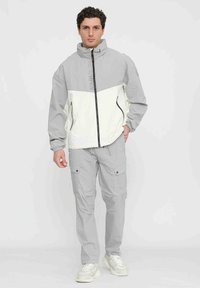 Giacca bi-colore grigia e bianca con colletto alto e zip nera, abbinata a pantaloni cargo grigi chiari. Presenta numerose tasche e una vestibilità rilassata.