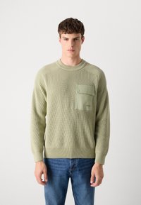 Pull en maille côtelée vert clair avec un col rond, doté d'une poche poitrine et d'un détail en tissu contrastant. Porté avec un jean en denim bleu.