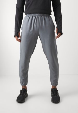 Person trägt graue Jogginghose mit elastischem Bund, schwarzen Langarmshirt mit Daumenlöchern und schwarze Sportschuhe, steht vor weißem Hintergrund.