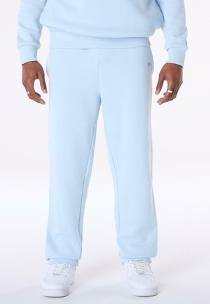 Person trägt hellblaue Jogginghose, passendes Sweatshirt, weiße Sneakers und silberne Ringe an beiden Händen, steht vor einem weißen Hintergrund.