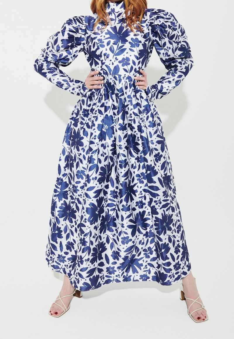 Femme portant une longue robe blanche à motif floral bleu avec des manches bouffantes, mains sur les hanches, debout, les pieds écartés, en talons beiges à brides.