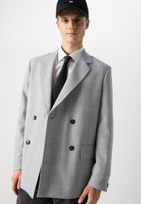 Gray plaid double-breasted blazer with notched lapels, black buttons, and a textured finish, worn over a white shirt and black tie.