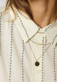 Chemise crème avec broderie de feuilles vertes le long de la patte de boutonnage, dotée d'un col classique et d'un collier en chaîne dorée avec un pendentif circulaire.