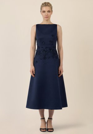 Frau mit blondem Haar in einem ärmellosen, marineblauen Midikleid mit Blumenstickerei und schwarzen Knöchelriemen-Absätzen, die vor einem beigen Hintergrund steht.