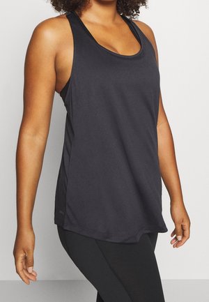 Kvinde iført en løs sort ærmeløs tanktop og sorte leggings, stående mod en ensartet lys baggrund.