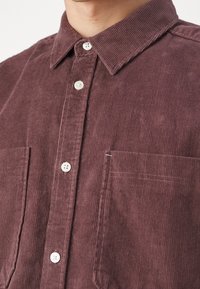 Camisa de pana burdeos oscuro con cuello clásico, placket de botones en la parte delantera, dos bolsillos en el pecho y tejido texturizado.