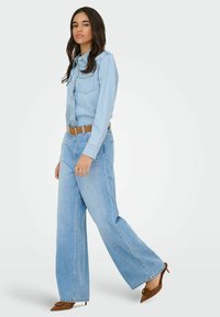 Jasnoniebieska koszula dżinsowa z zatrzaskami, zestawiona z luźnymi, jasnoniebieskimi dżinsami i brązowymi mule na obcasie. Faktura jest miękka, luźny krój.