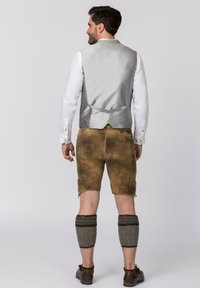 Grauer Satin-Weste über einem weißen Hemd, braune Shorts mit strukturierter Oberfläche und graue gestrickte Beinwärmer. Braune Schuhe ergänzen das Outfit.
