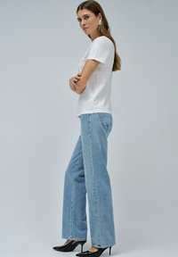 Camiseta blanca de manga corta, vaqueros anchos azules y tacones negros de punta. Tejido suave y líneas limpias. La modelo posa con los brazos cruzados.