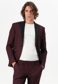 Burgunderfarbener Blazer mit schwarzen Satinrevers, einreihiger Knopfverschluss und passenden Hosen. Wird über einem weißen T-Shirt getragen und zeigt ein elegantes Design.