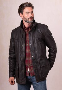 Bearded man wearing a dark brown leather jacket over a red plaid shirt and blue jeans, looking to the side against a plain background.