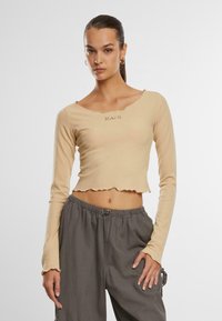 Rippstrukturierte, beigefarbene Crop-Top mit langen Ärmeln und gewelltem Saum. Verfügt über ein dezentes, gesticktes Logo auf der Vorderseite. Kombiniert mit grauen Hosen.