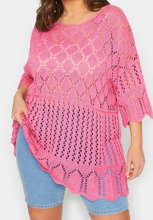 Femme portant un top asymétrique rose en maille ajourée sur un short en jean bleu clair, tenant le côté du top d'une main.