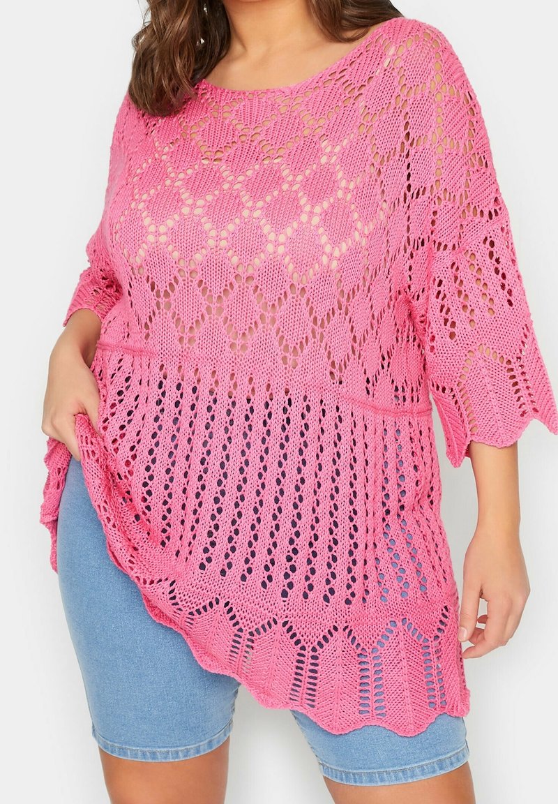 Femme portant un top asymétrique rose en maille ajourée sur un short en jean bleu clair, tenant le côté du top d'une main.