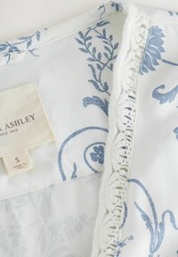 Baltas audinys su mėlynais gėlių raštais; turi tekstūrinį nėrinių apvadą. Etiketėje užrašyta "Laura Ashley", dydis S, pagaminta Indijoje.
