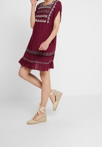 Burgundské šaty s krátkým rukávem, s vyšívanými detaily na těle a lemu, spárované s tanovými espadrilkami na platformě s vázáním kolem kotníku.