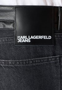 Crne traperice od denima s sjajnim crnim kožnim zaštitnim ušivenim detaljem sa utisnutim bijelim tekstom "KARL LAGERFELD JEANS." Uključuju vidljive šavove.