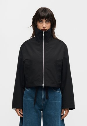 Άτομο με σκούρα μαλλιά φοράει μαύρο κοντό μπουφάν με ψηλό γιακά που έχει κλείσει εντελώς, σε συνδυασμό με άνετο μπλε τζιν, στέκεται μπροστά σε μονόχρωμο φόντο.