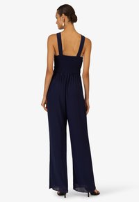 Navy blue jumpsuit featuring wide shoulder straps, a smocked bodice, and loose, fluid legs. Made from lightweight fabric with a soft texture.