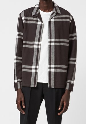 Muž v černobílé velké kostkované košili na zapínání přes bílým tričkem a černými kalhotami stojí před jednobarevným pozadím.