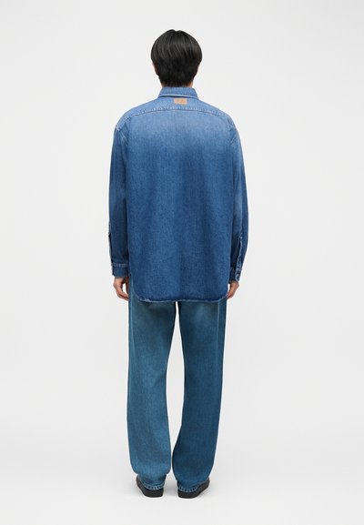 Personne debout, de dos, portant une chemise en denim bleu délavé et un jean bleu ample, sur un fond uni.