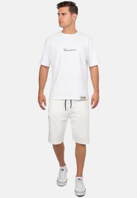 T-shirt in cotone bianco con testo in scrittura nera sul petto, abbinato a pantaloni corti bianchi con stampa del logo laterale. Completato con sneaker bianche.