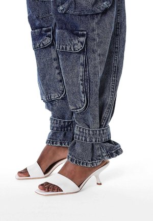 Džinsa garās bikses ar lielām kabatām un sadrupušām maliņām, kopā ar baltām atvērtām papēžu kurpēm ar elegantu dizainu un zemu papēdi.