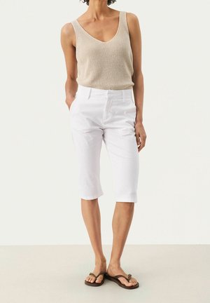Femme portant un haut en tricot sans manches beige, un short blanc longueur genou et des tongs marron foncé, debout avec une main dans la poche.