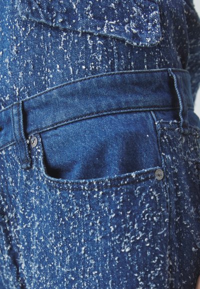 Denim jeans featuring a textured blue fabric with white splatters, a back pocket, and metal button accent at the waist.