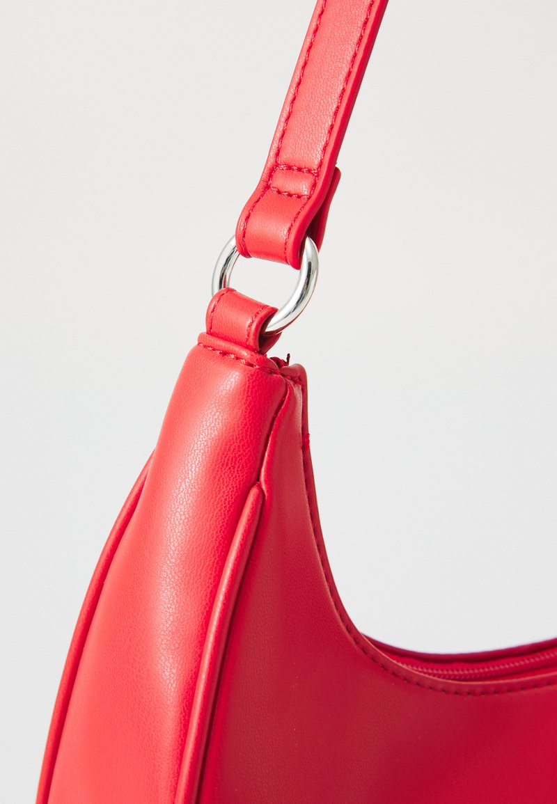 Bolso de hombro de cuero rojo con forma curvada, que presenta un anillo de metal plateado y una textura suave. Detalle de cremallera visible en la parte superior.