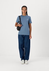 Camiseta de algodón azul con mangas cortas y un pequeño logo, combinada con jeans de mezclilla azul holgados y zapatillas blancas. Una bolsa de hombro cuelga de un hombro.