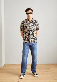 Schwarzes Kurzarmhemd mit floralen weißen Mustern, getragen mit blauen Jeans und schwarzen Sneakers. Das Model steht vor einem hellen Hintergrund.