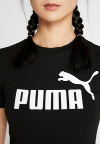 Kobieta z czarnymi zaplecionymi warkoczami, ubrana w czarny T-shirt z dużym białym logo Puma i napisem na piersi, na białym tle.