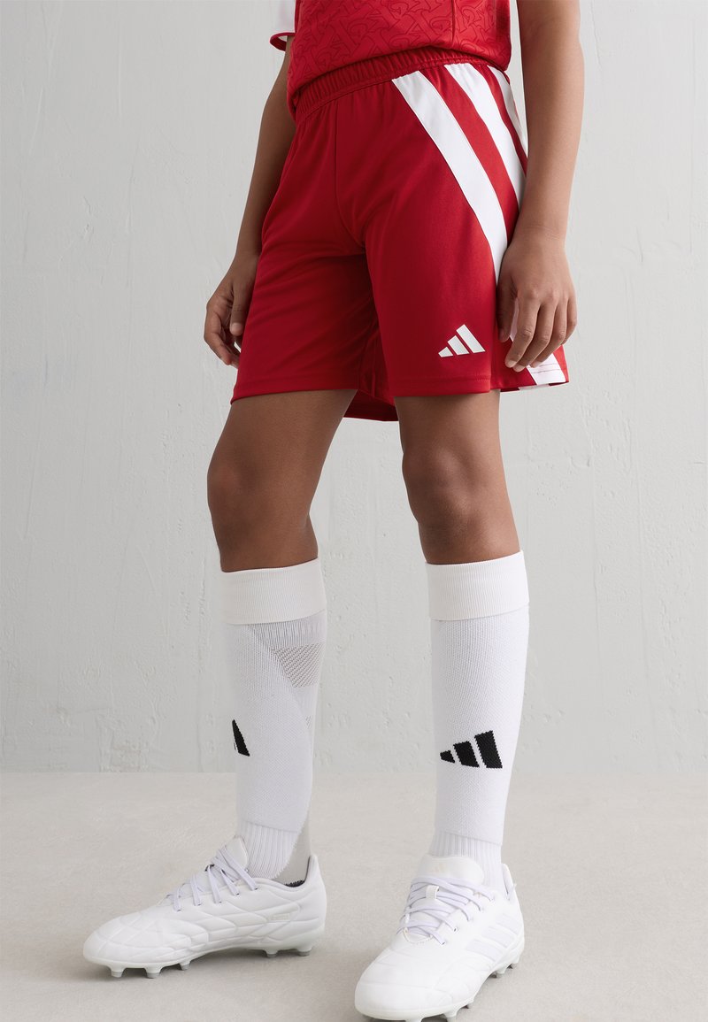 Rode sportieve shorts hebben witte diagonale strepen en een elastische tailleband. Gemaakt in combinatie met witte sokken en witte voetbalschoenen.