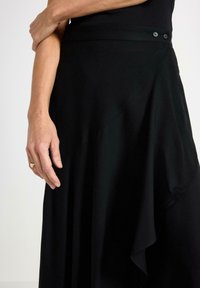 Kvinnas hand som vilar på en svart h�ögmidjad kjol med draperingar och knappar nära midjan mot en neutral bakgrund.