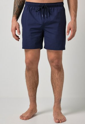 Mand iført marineblå badeshorts med snøre, synlige tatoveringer på venstre arm og højre ankel, stående barfodet på en lys baggrund.