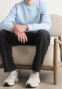 Sweatshirt bleu clair avec col rond et petit logo, associé à un jean noir et des baskets grises, assis sur une chaise à cadre en bois.