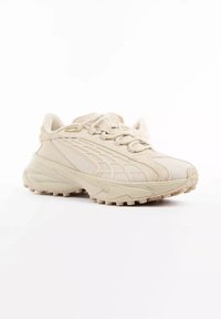 Baskets beige épaisses avec semelle texturée, panneaux en mesh et en daim, fermeture à lacets, présentées sous un léger angle sur fond blanc.