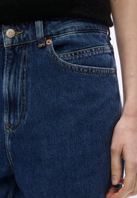 Mėlynos spalvos džinsai iš džinso audinio, su aukštu liemeniu, geltonomis siūlėmis, metaline sagtele ir priekiu kišene. Minkšta tekstūra su švelniu išblukimu.