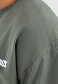 Camiseta de algodón verde con una textura suave, mostrando la costura del hombro y una parte del texto impreso en blanco en la parte inferior delantera.