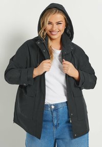 Svart huva jacka med framdragkedja, tryckknappar och stora fickor, tillverkad av ett slätt, matt tyg. Bärs över en vit skjorta och blå jeans.