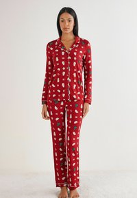 Červená pyžamová souprava se skládá z dlouhých ruk�ávů a kalhot, vyrobená z měkké látky, zdobená bílými a zelenými vzory s vánoční tématikou.