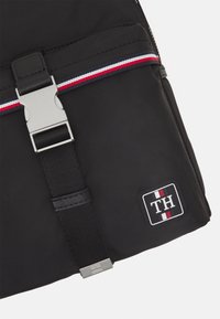 Zaino in nylon nero con un fermaglio argento, una striscia orizzontale rossa, bianca e blu, e una patch rettangolare con logo "TH".