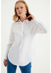 Witte lange mouwen blouse met een kraag, voorzien van een gebogen zoom en een gladde textuur, gecombineerd met blauwe strakke broek.