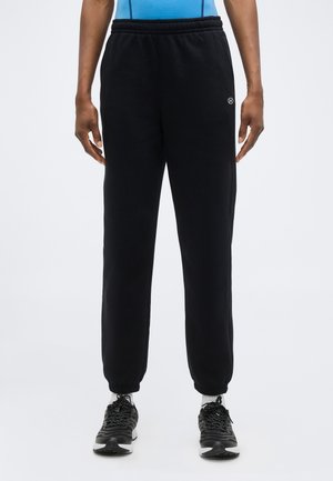 Mann trägt schwarze Sweatpants mit elastischem Bund und elastischen Bündchen, weiße Socken und schwarze Sportschuhe, steht vor einem einfarbigen Hintergrund.