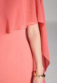 Korallfarbenes Kleid mit fließendem Chiffon-Überzug, der eine glatte Textur aufweist. Ein goldener Armreif schmückt das Handgelenk und betont das Design.