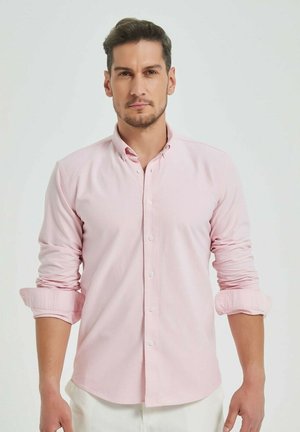 Chemise boutonnière rose clair avec un col classique, manches longues retroussées aux poignets, fabriquée dans un tissu doux avec une texture lisse.