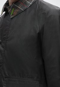 Gros plan d'une veste Barbour noire avec un grand bouton, un col à carreaux et des coutures visibles sur l'épaule et la poche.